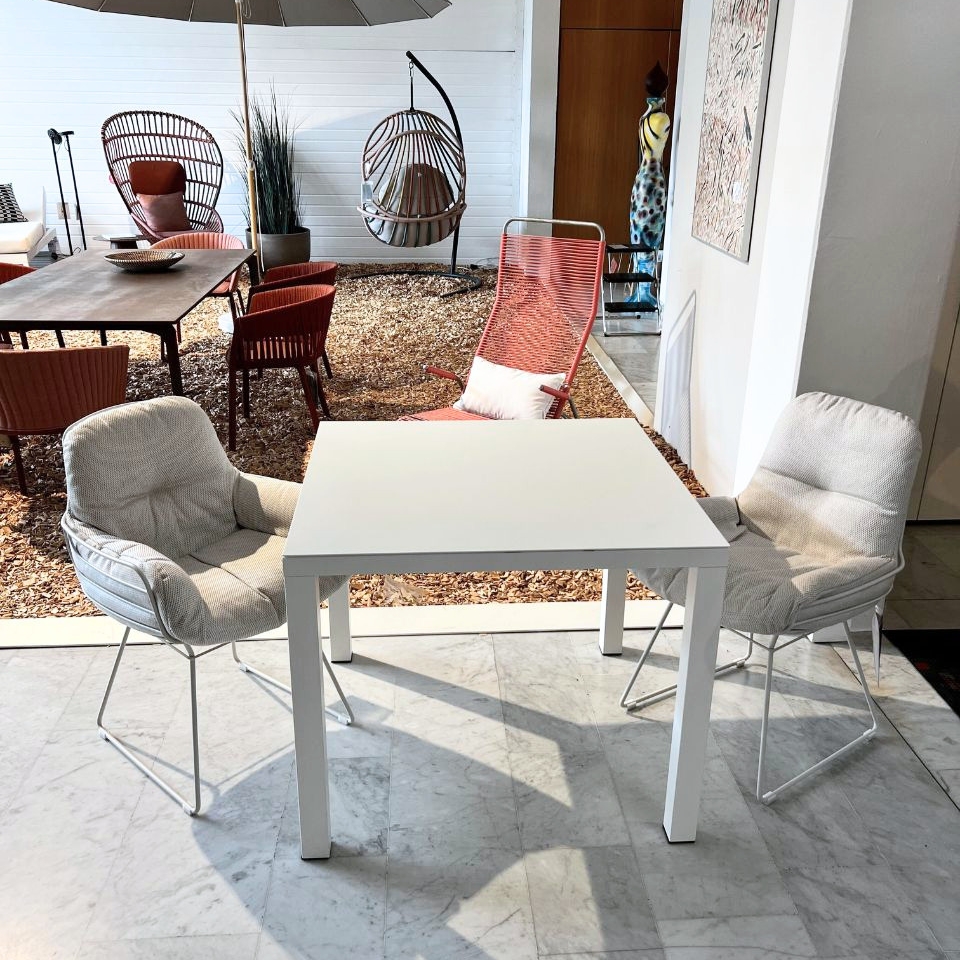White square table with two light gray cushioned chairs on a marble floor in a bright showroom; background shows outdoor patio seating with red chairs and hanging wicker chairs under an umbrella.