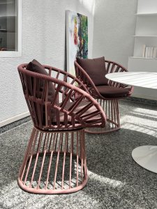 Two pink circular rope chairs with dark cushions sit beside a white round table in a sunlit indoor seating area, with an abstract painting leaning against the wall.