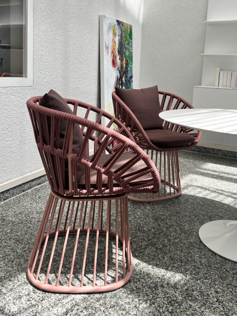 Two pink circular rope chairs with dark cushions sit beside a white round table in a sunlit indoor seating area, with an abstract painting leaning against the wall.