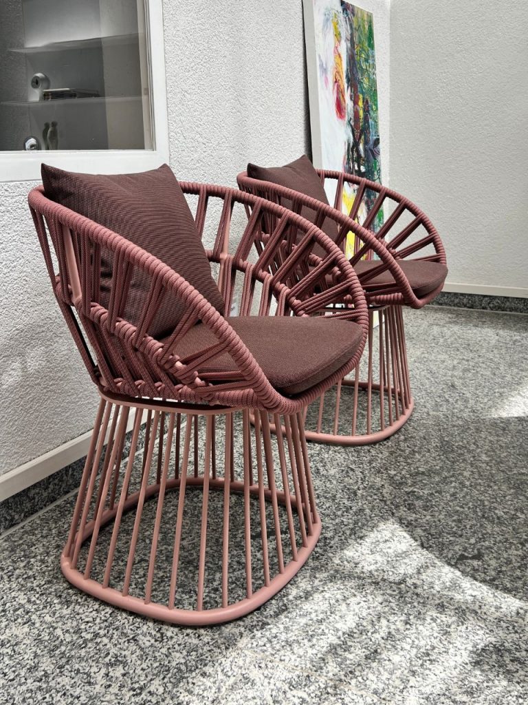 Two pink woven circular chairs with brown cushions in a white-walled hallway, with a colorful abstract painting beside them.