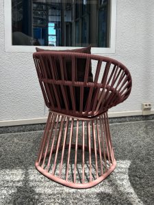 Pink circular patio chair with vertical spokes and a cushion on a gray tiled floor beside a white wall and window.