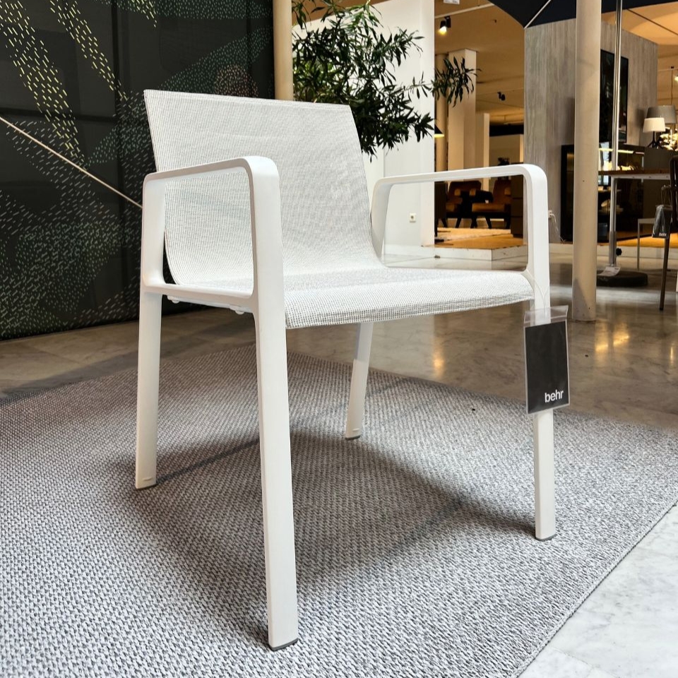 White modern armchair with a woven light-gray seat and back, on a gray rug in a showroom; Behr tag hangs from the front leg.