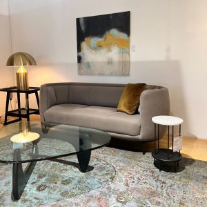 Modern living room scene with a gray curved sofa, mustard pillow, glass coffee table, and abstract painting above the sofa, in a showroom setting.