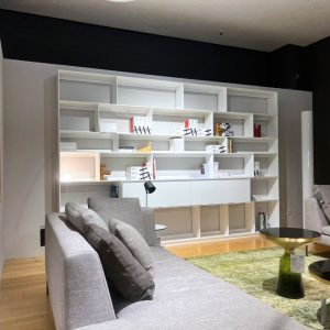 Modern living room showroom scene with a large white built-in shelving unit, grey sofa with cushions, black task lamp, and two round glass-top coffee tables on a green rug.