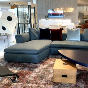 Blue sectional sofa with multicolored cushions in a modern furniture showroom, with a wooden coffee table and a patterned rug in the foreground.