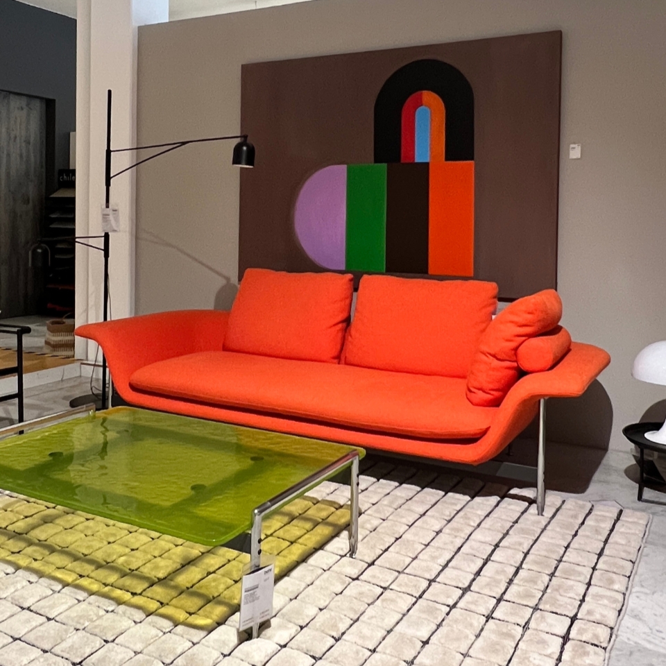 Bright orange modern sofa with matching cushions in a showroom, a large abstract painting on the wall, and a lime-green glass coffee table on a beige patterned rug