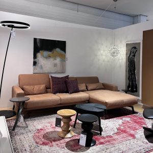 Modern living room showroom with a tan leather sectional sofa, accent pillows, abstract wall art, and a geometric pendant light in a bright space.