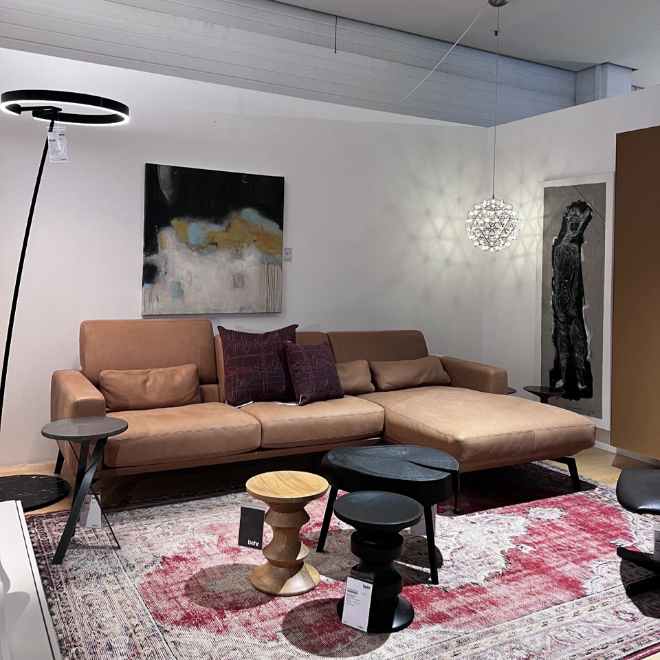 Modern living room showroom with a tan leather sectional sofa, accent pillows, abstract wall art, and a geometric pendant light in a bright space.