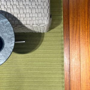 Wicker chair on a green striped outdoor rug beside a round dark table, with sunlit wooden floor planks nearby.