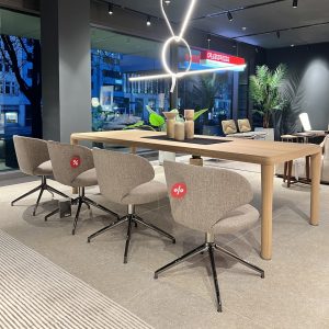 A long wooden table with four beige swivel chairs in a showroom, modern pendant rings overhead and large windows in the background.