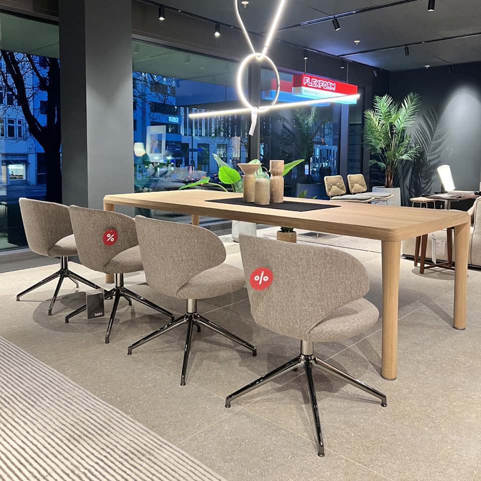 A long wooden table with four beige swivel chairs in a showroom, modern pendant rings overhead and large windows in the background.