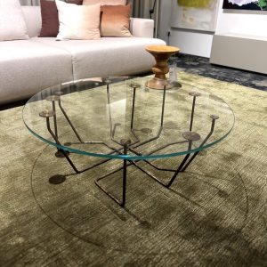Round glass coffee table with a sculptural metal base in a living room showroom, beige sofa and cushions in the background, and a wooden stool on top of the table.