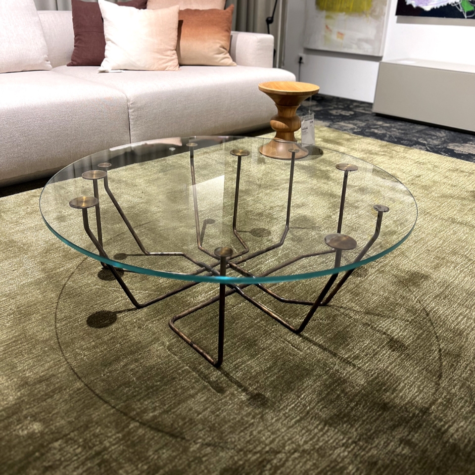 Round glass coffee table with a sculptural metal base in a living room showroom, beige sofa and cushions in the background, and a wooden stool on top of the table.