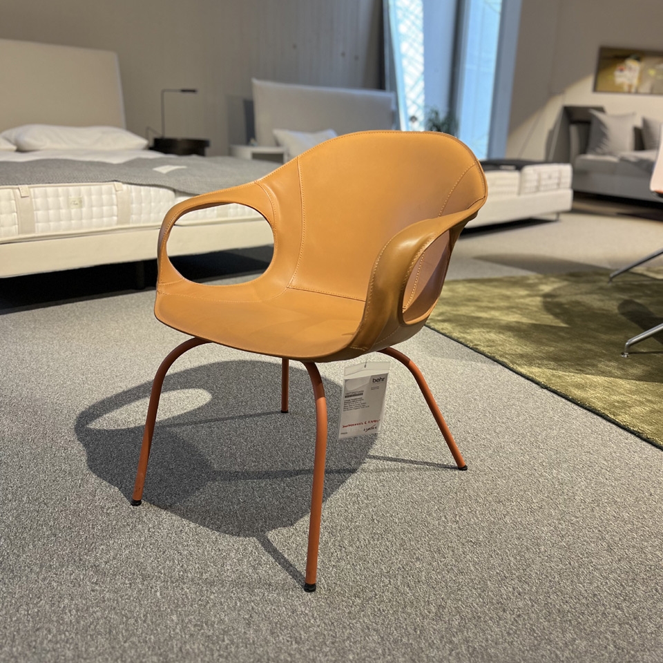 Tan leather lounge chair with curved back and cutout armrests, on orange metal legs in a showroom setting with a bed and sofa in the background.