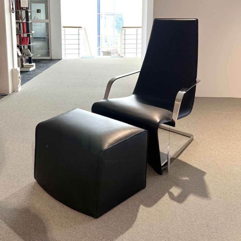 Black leather lounge chair with a matching square ottoman in a modern office space on a beige carpet.