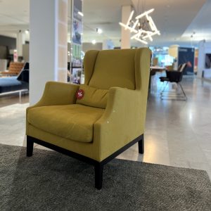 Mustard-yellow upholstered armchair with high back and curved arms, placed on a dark rug in a showroom. A red '%' sale tag rests on the back cushion.
