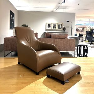 Brown leather lounge chair with a matching ottoman displayed on a wooden showroom floor.
