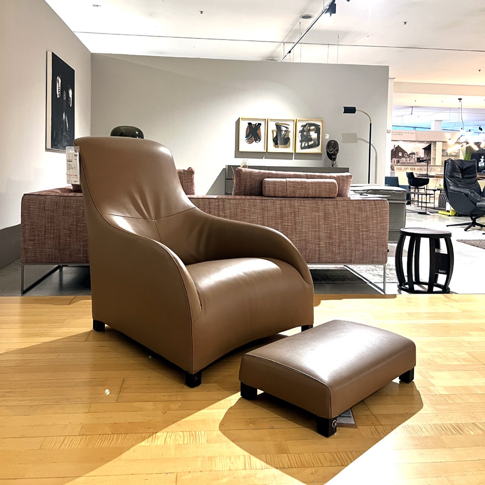 Brown leather lounge chair with a matching ottoman displayed on a wooden showroom floor.