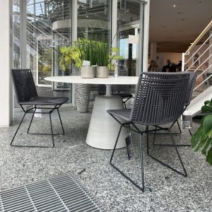 Outdoor showroom seating: round white table with four black woven chairs around it, potted plants on the table, glass storefront behind.