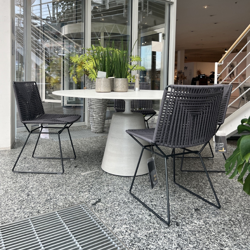 Outdoor showroom seating: round white table with four black woven chairs around it, potted plants on the table, glass storefront behind.
