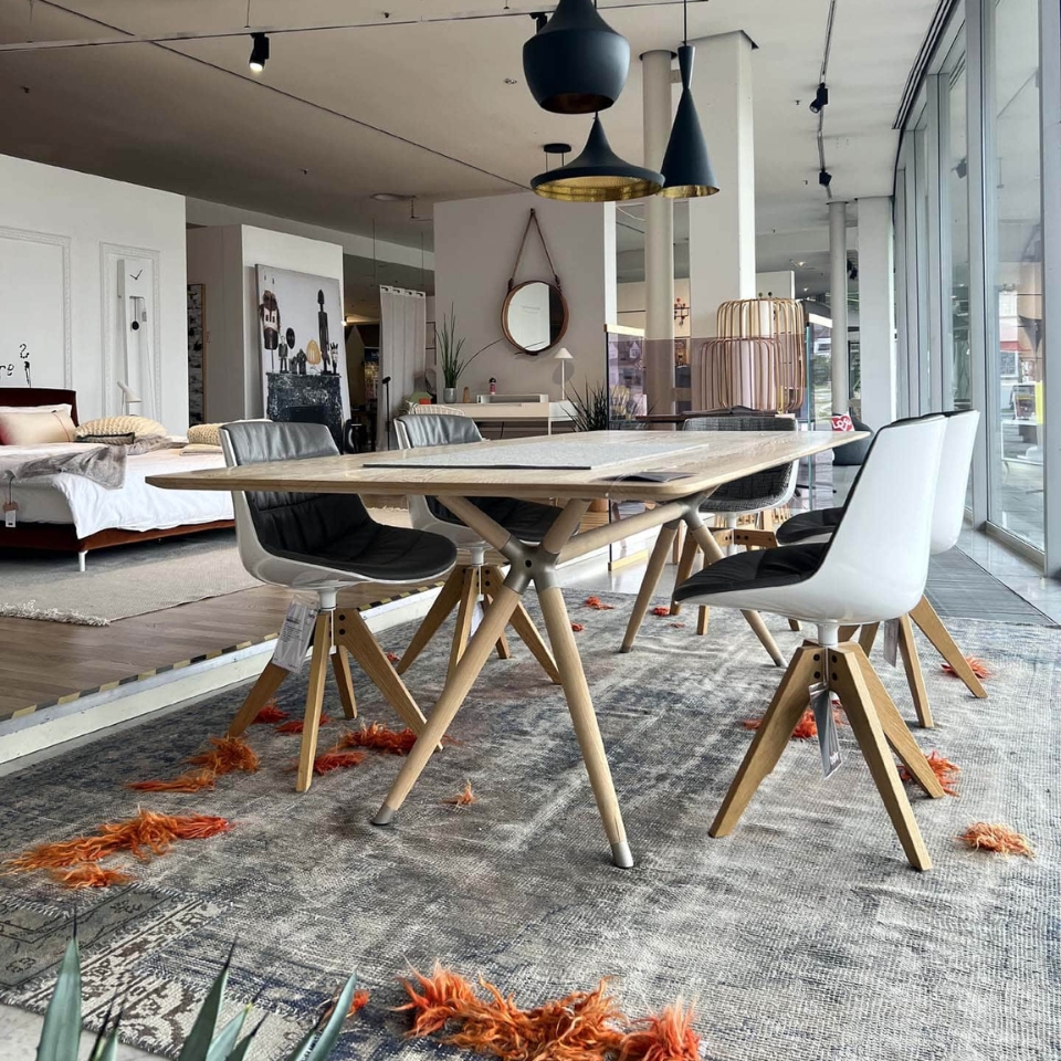 Open-plan furniture showroom with a light wood dining table and white/black chairs on a large patterned rug, pendant lights overhead, and beds and decor in the background.