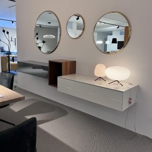 Interior showroom scene: wall with three gold-framed round mirrors above a white floating cabinet displaying two decorative lamps and a small wooden box edge-to-wall.