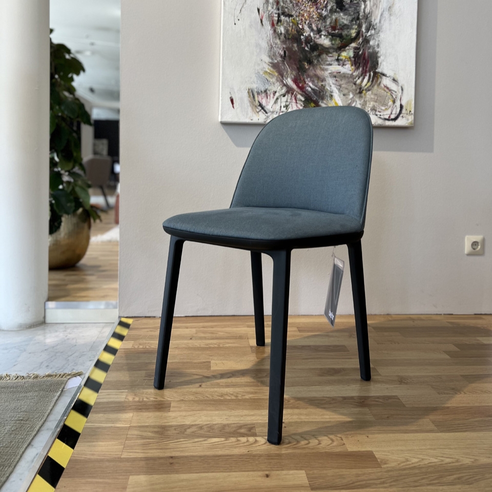 Blue upholstered chair with black legs in a showroom, tag hanging from the side, against a light wall with abstract painting above it.