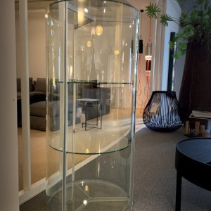 Tall cylindrical glass display cabinet with two shelves in a modern showroom, reflecting lights and surrounding furniture.