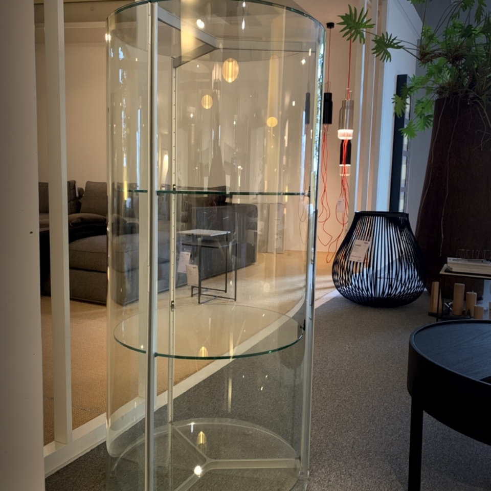 Tall cylindrical glass display cabinet with two shelves in a modern showroom, reflecting lights and surrounding furniture.