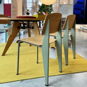 Set of light wood chairs with mint-green legs around a wooden table on a yellow rug in a showroom. Tags hang from chairs.