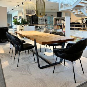 Wooden rectangular table surrounded by six black leather chairs in a bright, modern showroom with pendant lamps overhead and a staircase in the background.