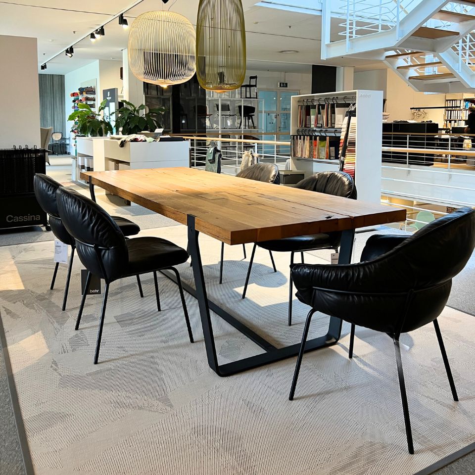 Wooden rectangular table surrounded by six black leather chairs in a bright, modern showroom with pendant lamps overhead and a staircase in the background.