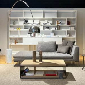 Modern living room setup: gray sectional sofa with matching cushions, a tall arched metallic floor lamp, and a large white bookshelf filled with books and decorative items behind a light wood coffee table on a beige rug.