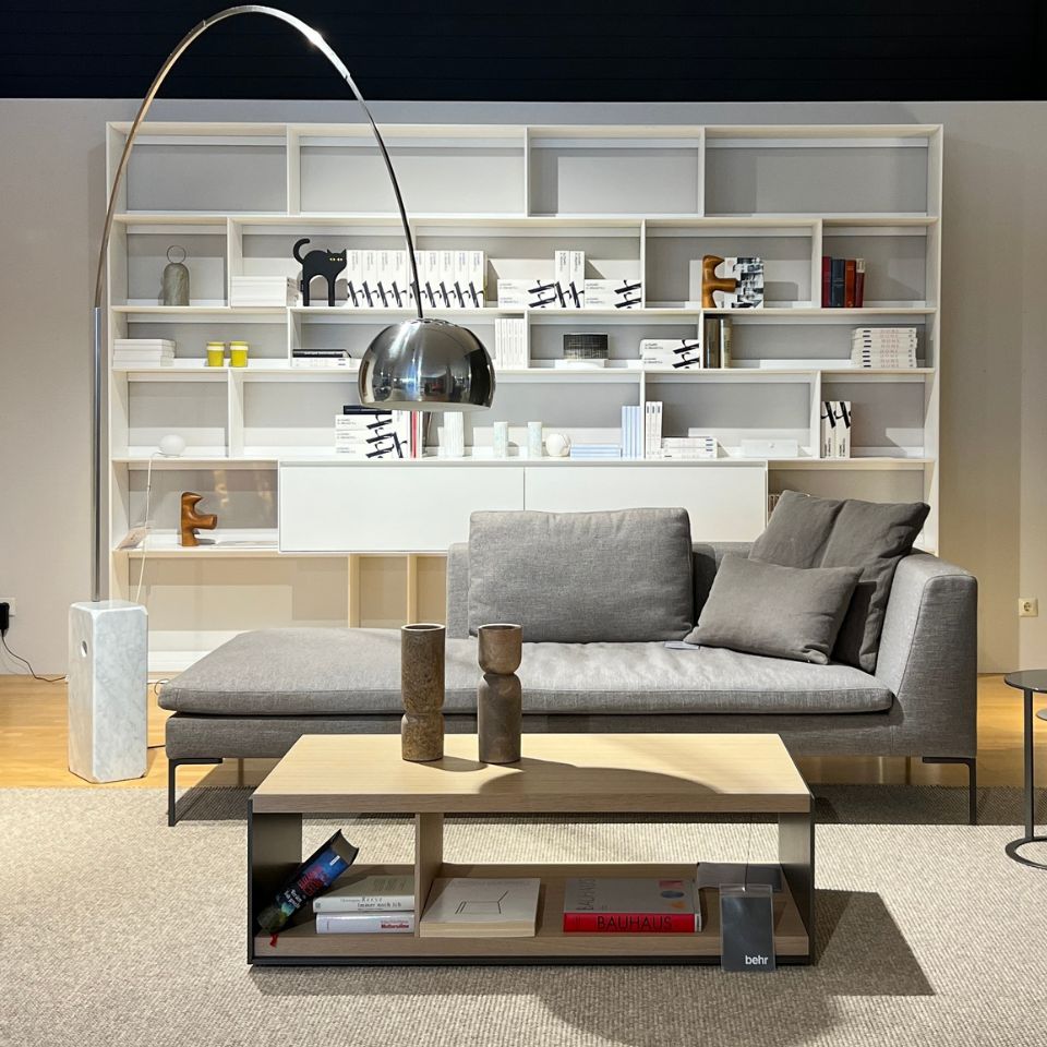 Modern living room setup: gray sectional sofa with matching cushions, a tall arched metallic floor lamp, and a large white bookshelf filled with books and decorative items behind a light wood coffee table on a beige rug.
