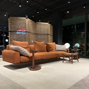 Orange fabric sofa with multiple cushions in a modern showroom, flanked by round wooden tables and a woven room divider behind.
