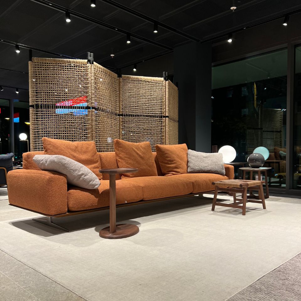 Orange fabric sofa with multiple cushions in a modern showroom, flanked by round wooden tables and a woven room divider behind.