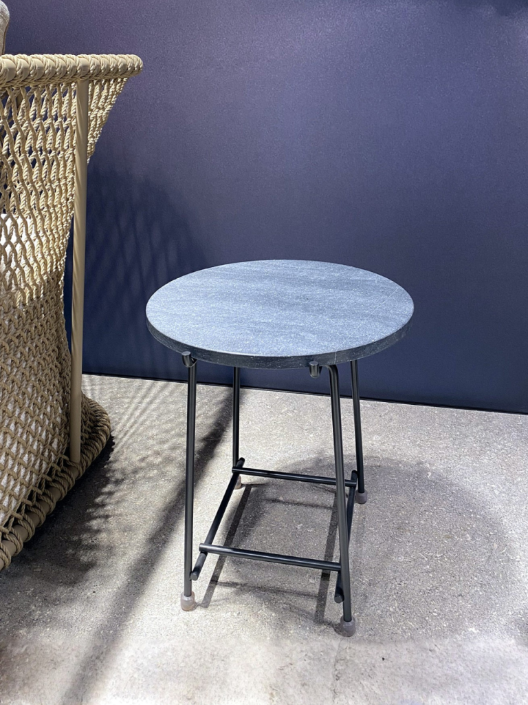 Small round blue-gray table on a black metal frame placed outdoors beside a woven wicker chair against a dark blue wall.