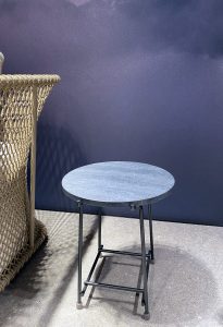 Wicker chair beside a round blue-speckled metal table against a dark blue wall outdoors.
