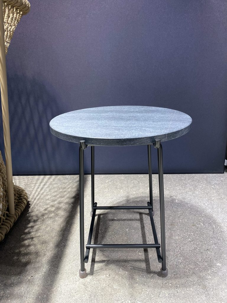 Round speckled gray granite tabletop on tall black metal legs, outdoors against a dark blue wall with rope coiled on the left.