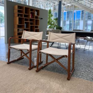 Two beige fabric-covered folding chairs with wooden frames sit in a bright, modern lobby beside a tall wooden bookshelf.