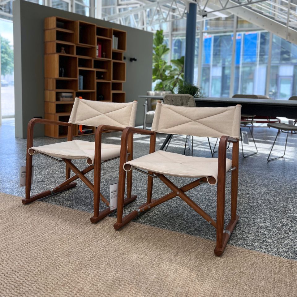 Two beige fabric-covered folding chairs with wooden frames sit in a bright, modern lobby beside a tall wooden bookshelf.