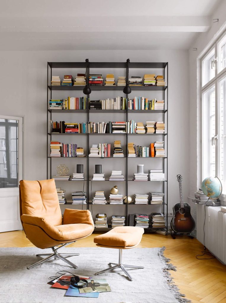 Living room with a tall black bookshelf full of books, two orange lounge chairs and an ottoman on a gray rug, guitar by the window.