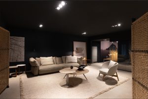 Contemporary living room showroom: beige sofa, round coffee tables, and a white upholstered chair on a textured rug in a dim, dark-walled space.