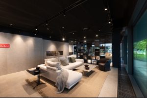 Modern showroom living area: white sectional sofa with cushions, gray throw, and a coffee table on a beige rug.