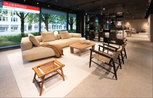 Modern showroom living area with a beige sectional sofa, wooden coffee table, and woven chairs under ambient track lighting by large windows.
