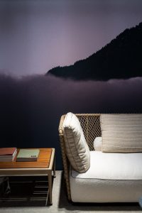 Wicker sofa with beige cushions sits beside a wooden coffee table holding books, against a dark purple mountain mural backdrop.