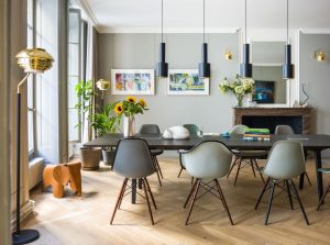 Bright dining room with a long dark table, mid-century chairs, and black pendant lights over a sunlit space with plants and art on the gray walls