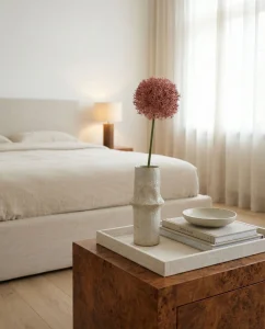 Neutral bedroom scene with a textured vase holding a pink spherical allium on a stack of books atop a wooden nightstand