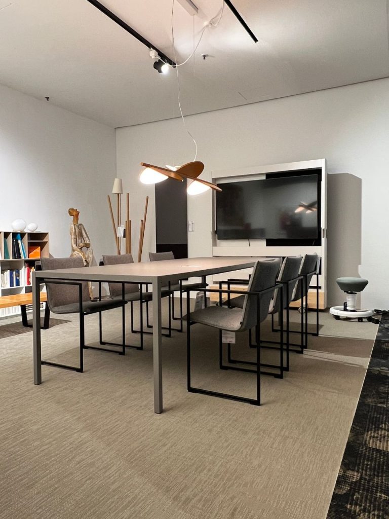Modern conference room with a long rectangular table, eight gray chairs, and a wall-mounted flat-screen TV on the far wall.