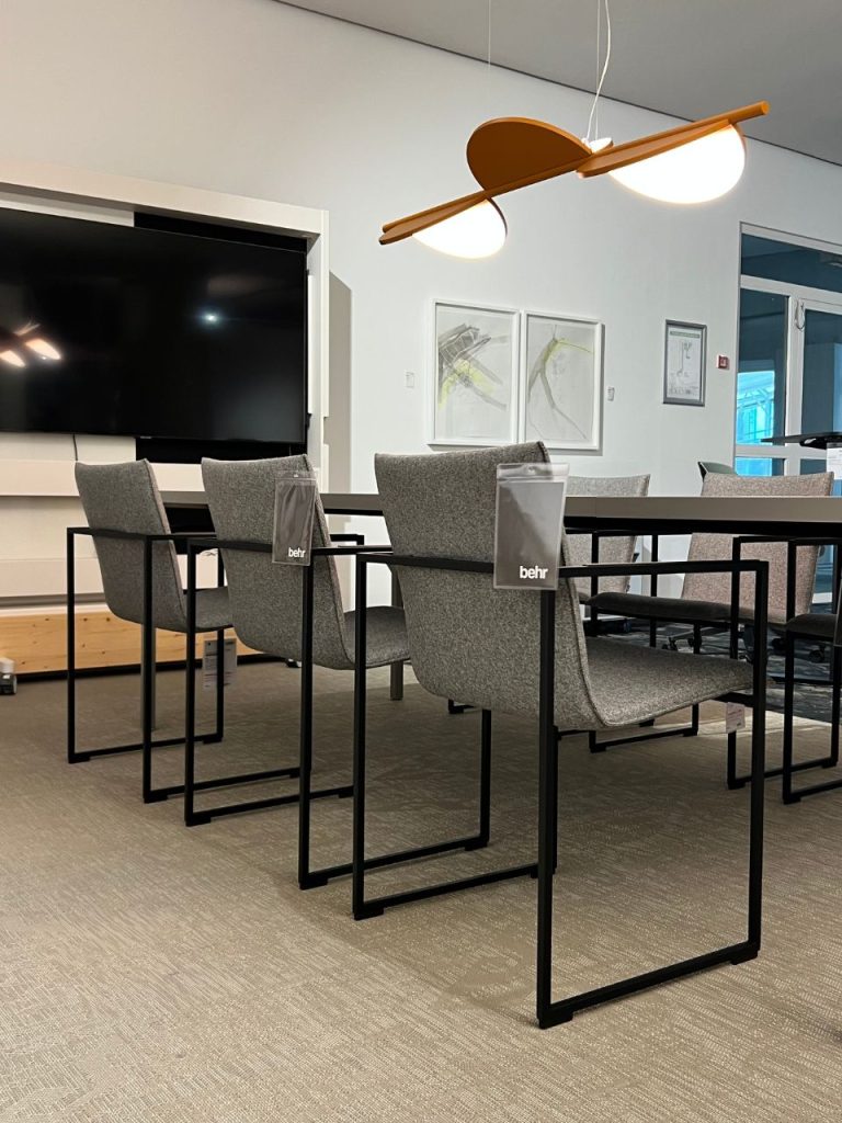 Conference room with gray upholstered chairs and black metal frames around a long table, a large wall-mounted TV, and a modern pendant light overhead.